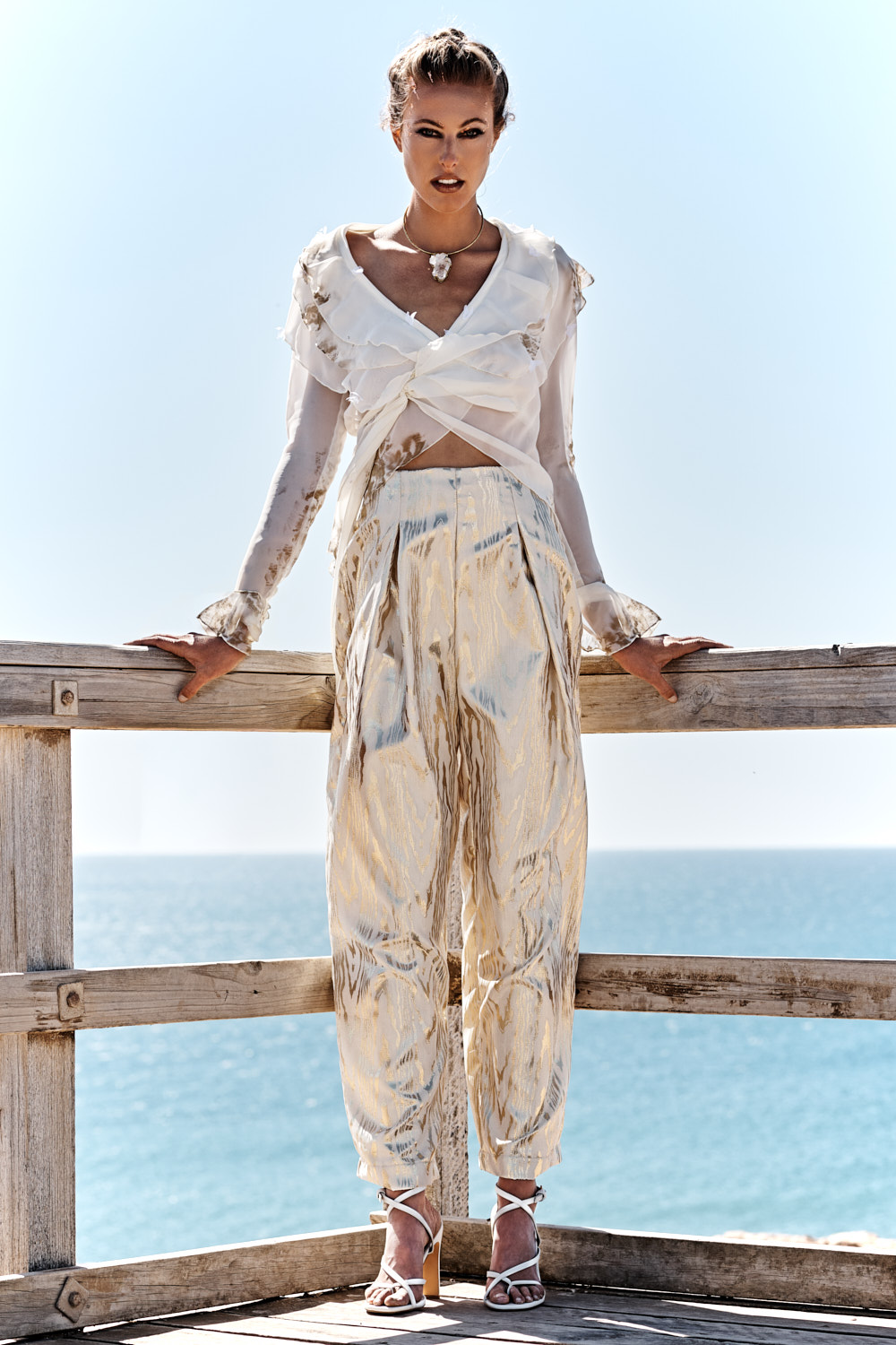 Fashion Photography Adelaide. A woman stands on a wooden deck overlooking the ocean, wearing a sheer white wrap top and high-waisted, shimmery gold pants, with white strappy heels. Her hair is styled up and she poses confidently against the railing.
