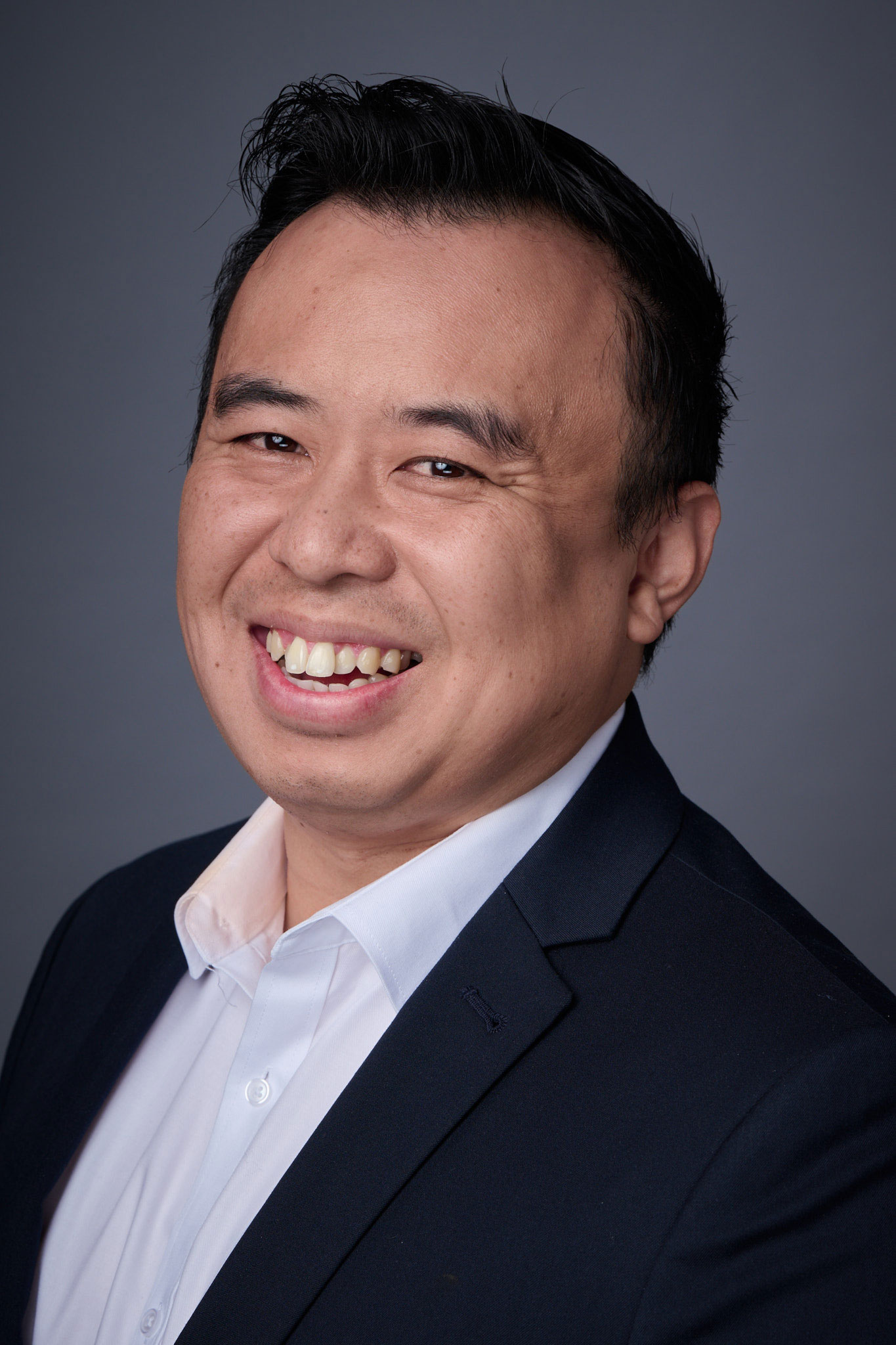 A man in a dark suit and white shirt smiles warmly at the camera against a plain gray background.