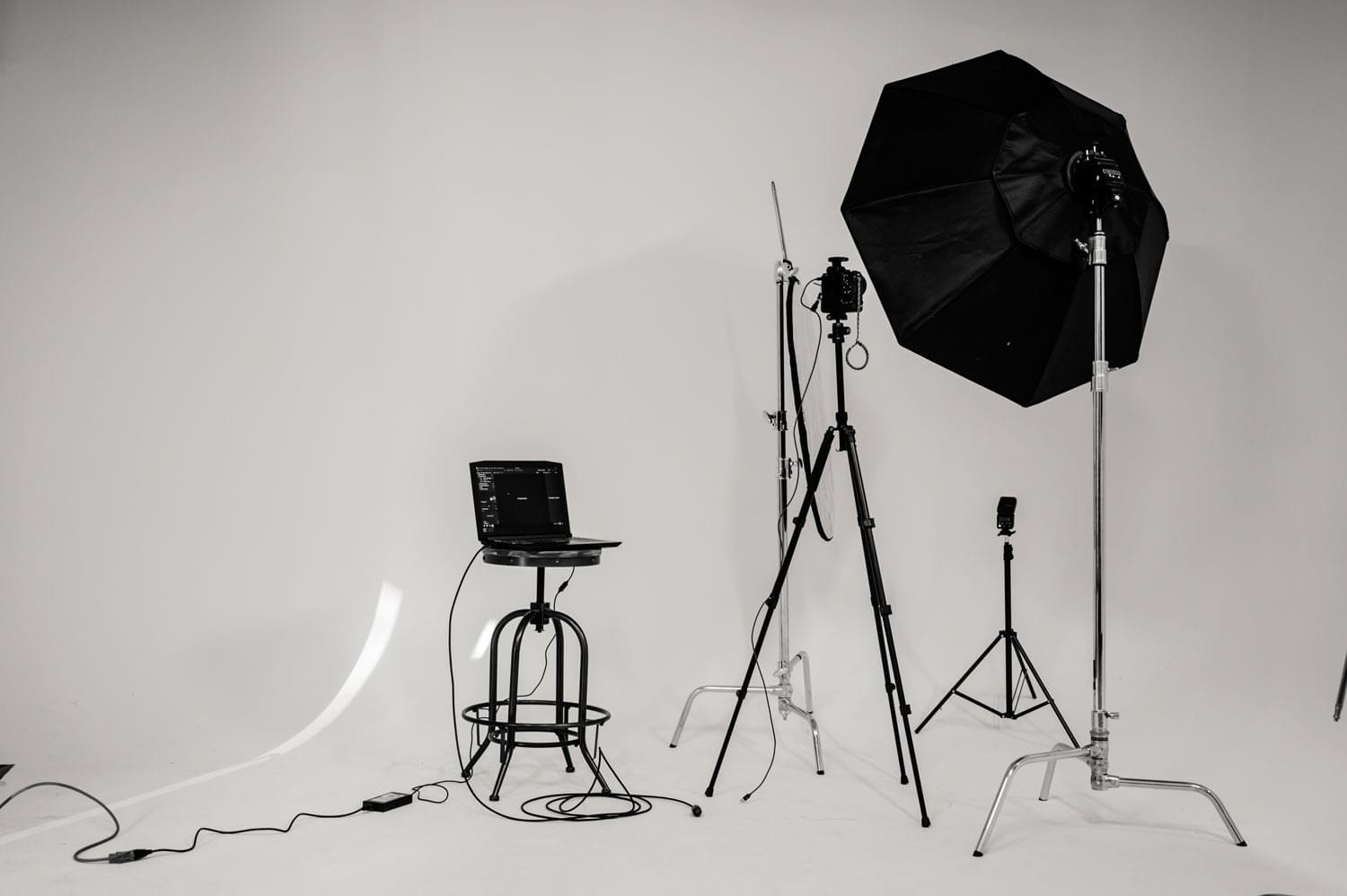 A photography studio setup with a laptop on a stool, a camera on a tripod, a large umbrella light, and various lighting stands and cables against a plain white background.