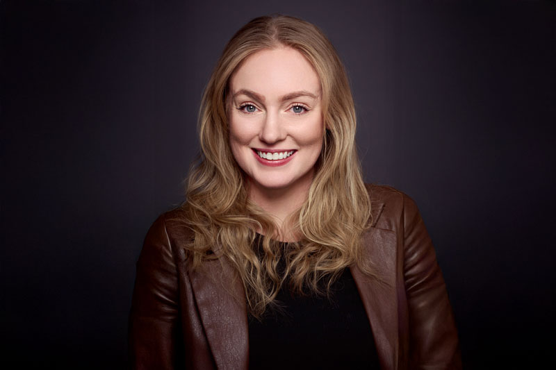 A woman with long, wavy blonde hair smiles at the camera. She is wearing a brown leather jacket over a black top and is posed against a dark background.